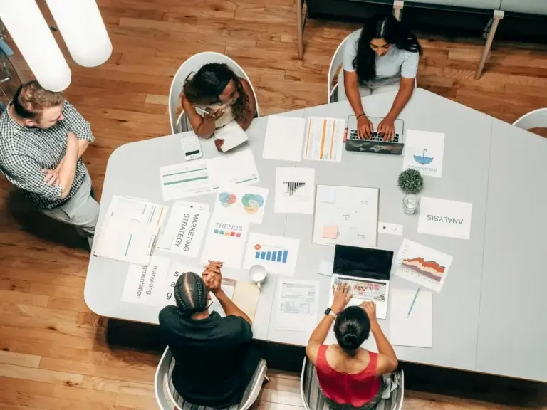 Aerial view of five people collaborating at a table covered with graphs, charts, and documents. One person is typing on a laptop, while others are reading or discussing the materials. The setting is a modern, brightly lit office with wooden floors.