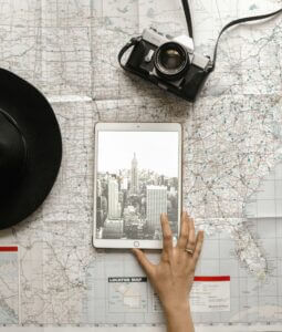 A tablet displaying a black and white photo of the Empire State Building rests on an open map. A hand reaches towards the tablet. A black hat and a vintage camera are placed nearby on the map, creating a travel-themed arrangement.