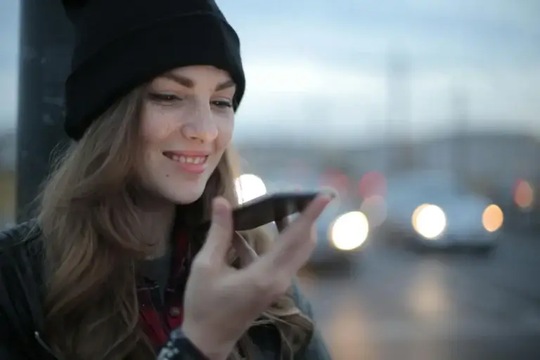 A young woman with long hair, wearing a black beanie and plaid shirt, smiles while holding a smartphone close to her face. She appears to be using the voice feature. The background is blurred with vehicle headlights on a street during dusk.