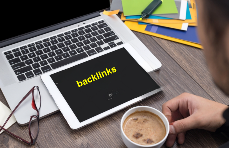 A person holds a cup of coffee in front of a laptop and a tablet with the word "backlinks" displayed on the screen. The scene also includes scattered papers, folders, and a pair of glasses on a wooden desk.