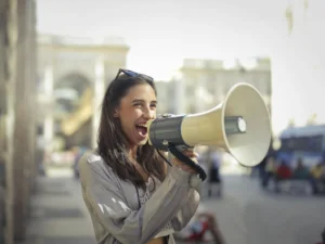 Woman with long hair and sunglasses on head, wearing a light jacket, standing in an outdoor urban area. She is holding and speaking into a large megaphone, smiling with an open mouth. Blurred background of buildings and people.